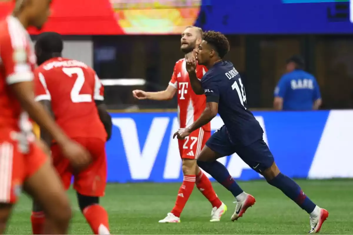 A player in a blue uniform celebrates a goal by making a silence gesture while several players in red uniforms walk on the soccer field. A player in a blue uniform celebrates a goal by making a silence gesture while several players in red uniforms walk on the soccer field.
