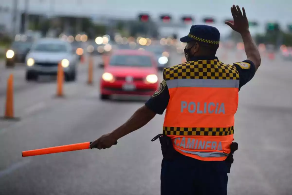 A traffic police officer wearing an orange vest is directing traffic on an avenue with several cars and orange cones. A traffic police officer wearing an orange vest is directing traffic on an avenue with several cars and orange cones.