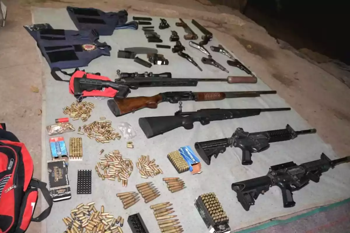 Several long and short firearms, ammunition, magazines, bulletproof vests, and backpacks are arranged on a blanket on the floor. Several long and short firearms, ammunition, magazines, bulletproof vests, and backpacks are arranged on a blanket on the floor.
