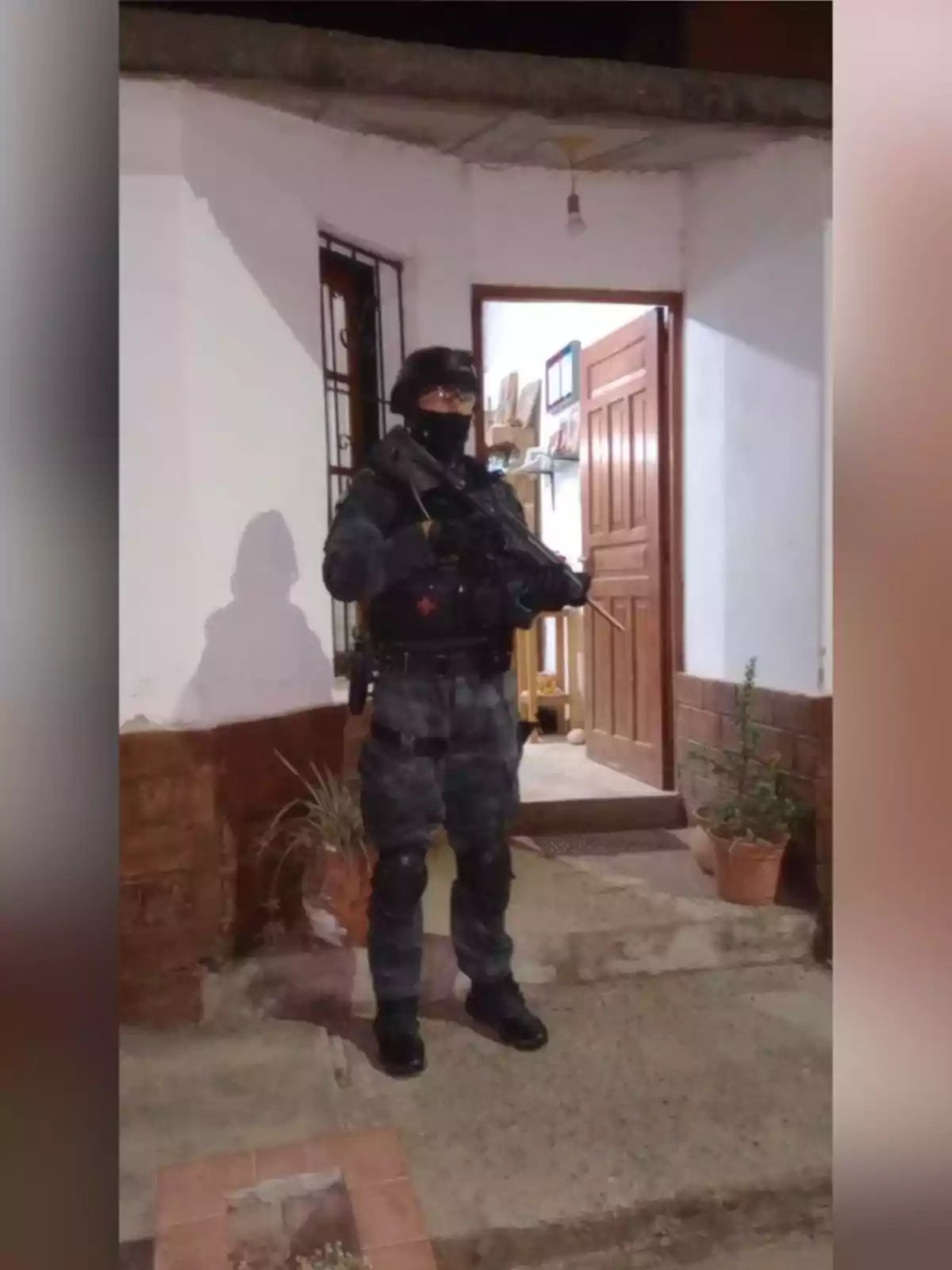 Persona con uniforme táctico y casco de protección sosteniendo un arma frente a la entrada de una casa blanca con plantas en macetas Persona con uniforme táctico y casco de protección sosteniendo un arma frente a la entrada de una casa blanca con plantas en macetas