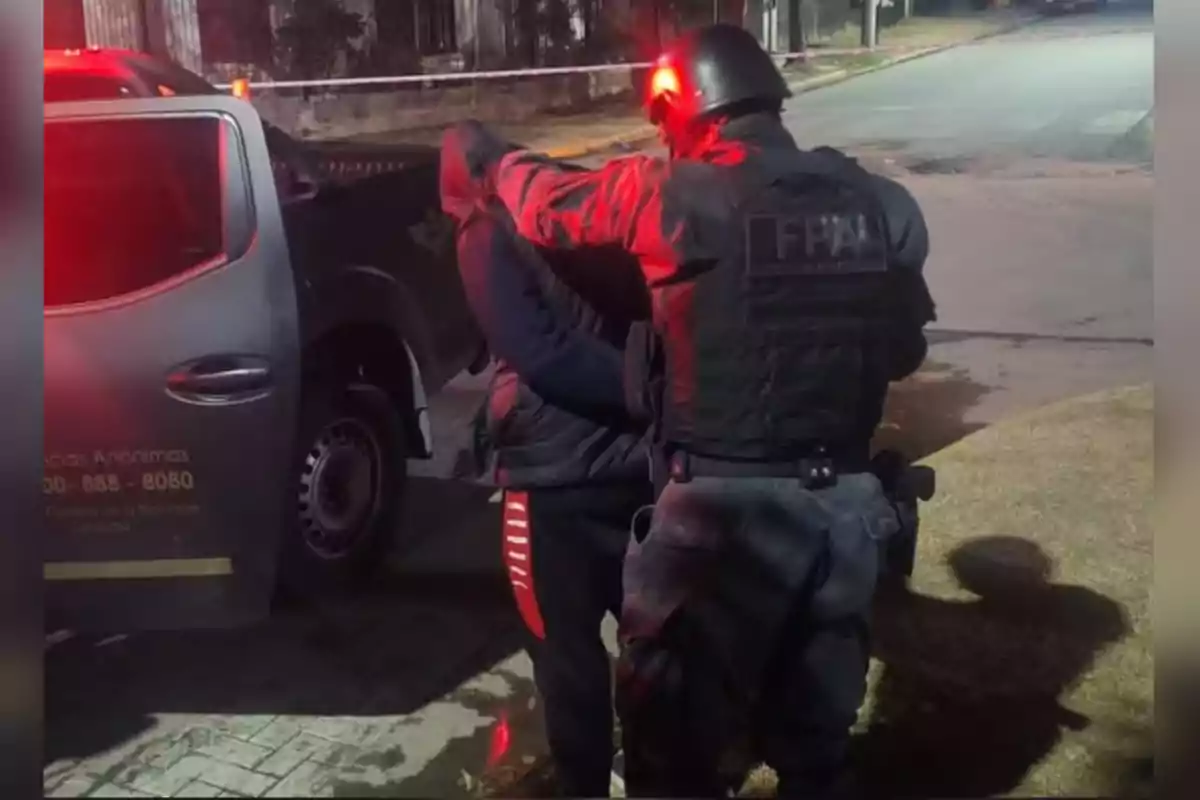 Un policía con chaleco y casco detiene a una persona junto a una camioneta en una calle por la noche. Un policía con chaleco y casco detiene a una persona junto a una camioneta en una calle por la noche.