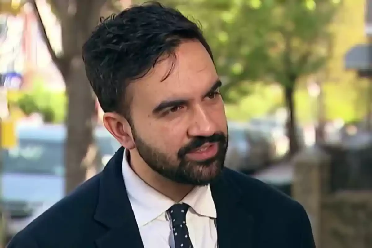 Hombre de cabello oscuro y barba con traje y corbata hablando al aire libre en un entorno urbano con árboles y autos al fondo