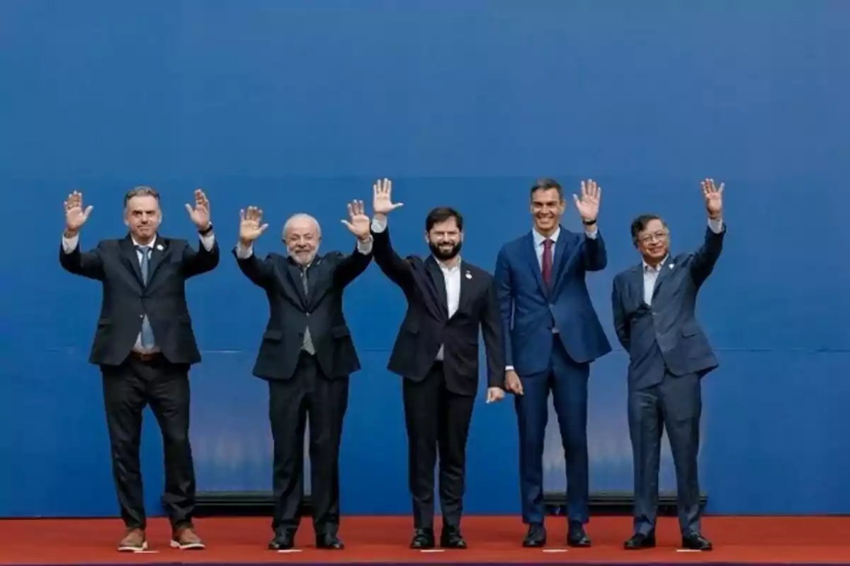 Cinco hombres con traje posan de pie sobre una alfombra roja frente a un fondo azul, levantando la mano en señal de saludo. Cinco hombres con traje posan de pie sobre una alfombra roja frente a un fondo azul, levantando la mano en señal de saludo.