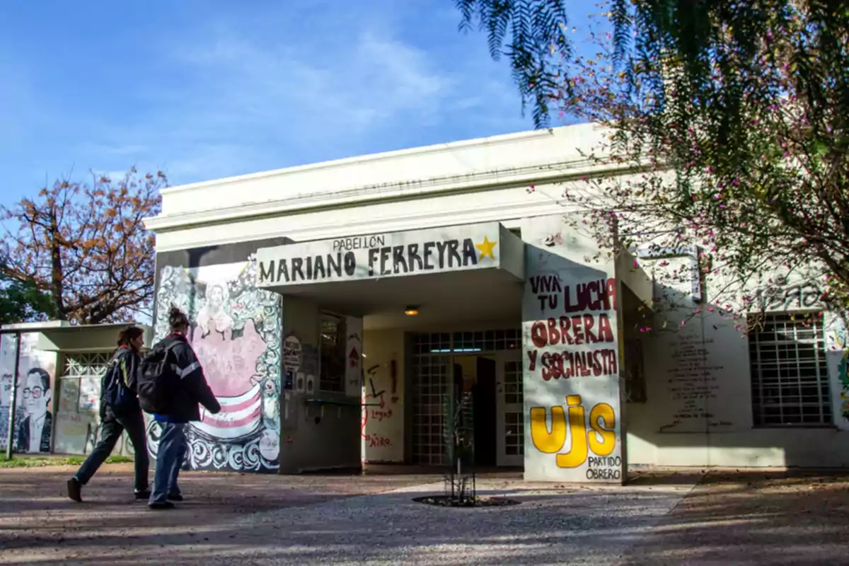 Dos personas caminan frente a un edificio con murales y grafitis en la entrada que tiene un cartel que dice Pabellón Mariano Ferreyra Dos personas caminan frente a un edificio con murales y grafitis en la entrada que tiene un cartel que dice Pabellón Mariano Ferreyra