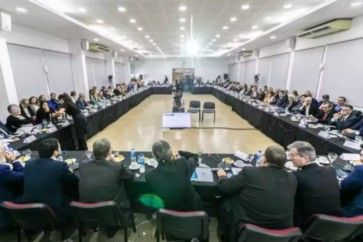 Personas reunidas en una gran sala de conferencias sentadas alrededor de mesas en forma de U participando en una reunión formal Personas reunidas en una gran sala de conferencias sentadas alrededor de mesas en forma de U participando en una reunión formal