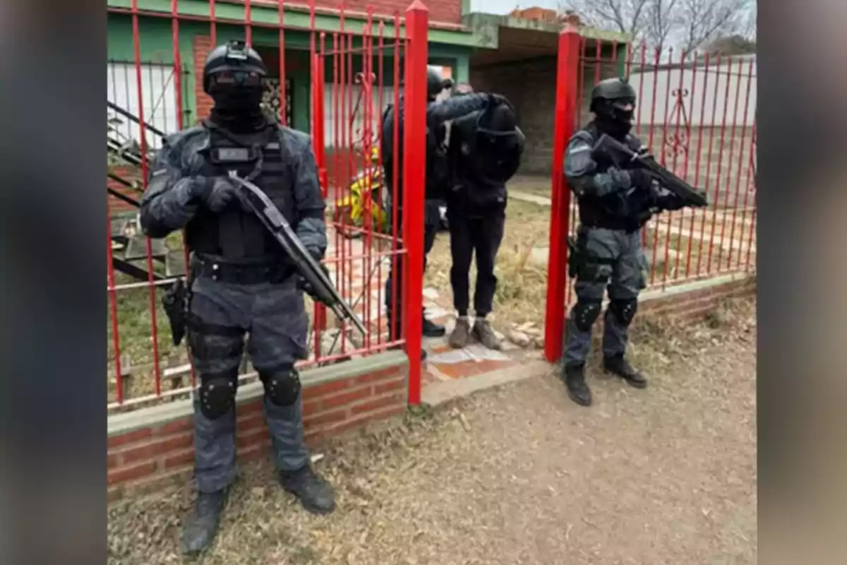 Three heavily armed police officers dressed in tactical gear guard a detained person in front of a house with red bars. Three heavily armed police officers dressed in tactical gear guard a detained person in front of a house with red bars.