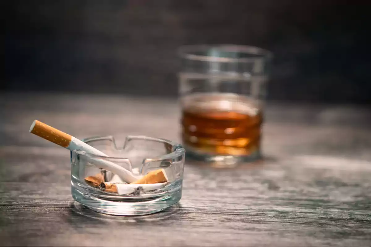 Cigarrillo apagado en un cenicero de vidrio con un vaso de whisky al fondo sobre una mesa de madera