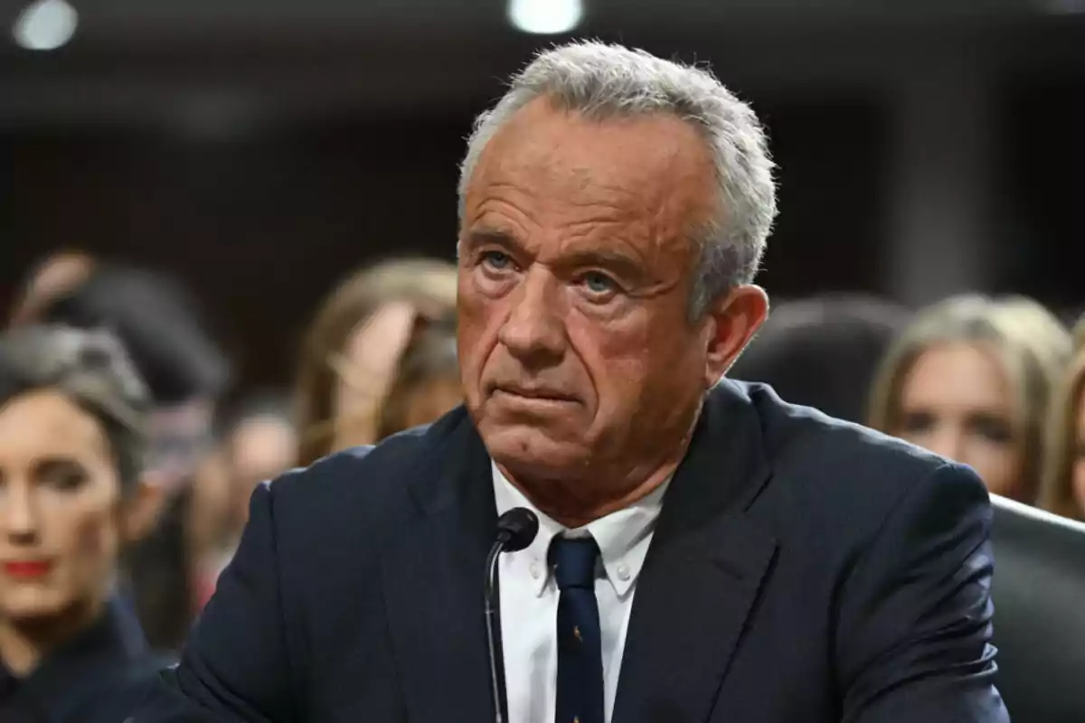 Hombre de cabello canoso con expresión seria usando traje oscuro y corbata sentado frente a un micrófono en un evento formal con personas al fondo desenfocadas Hombre de cabello canoso con expresión seria usando traje oscuro y corbata sentado frente a un micrófono en un evento formal con personas al fondo desenfocadas