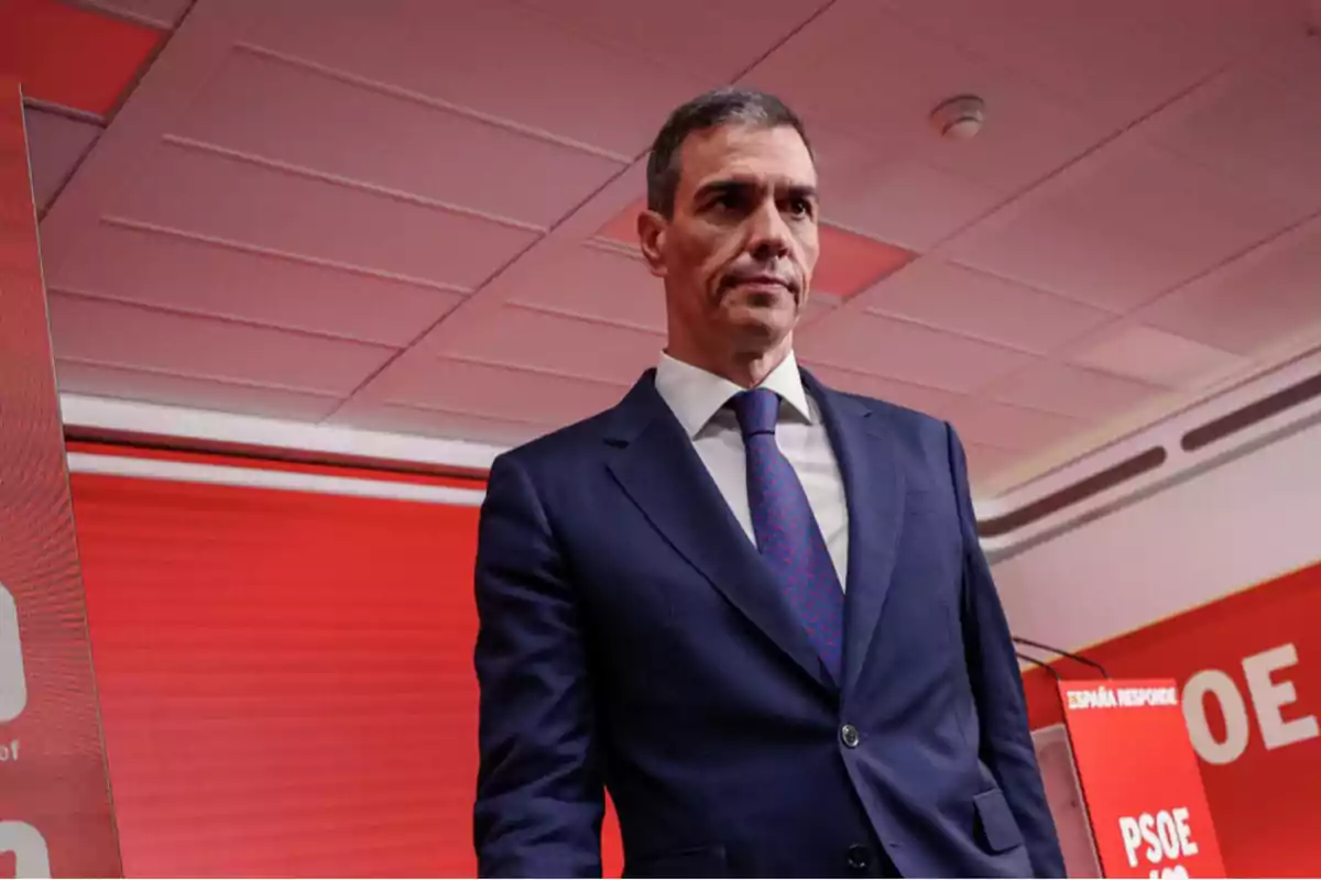 Hombre con traje azul y corbata de pie en un escenario con fondo rojo y letreros de PSOE