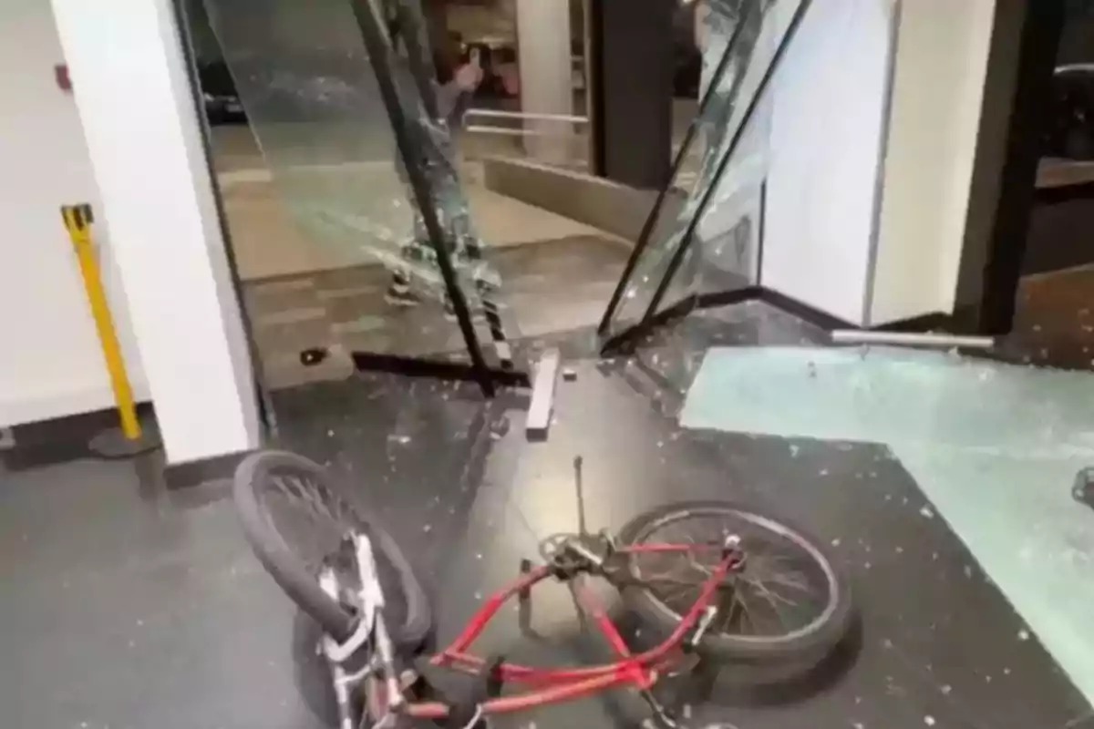Una bicicleta roja tirada en el suelo frente a una puerta de vidrio rota con fragmentos de cristal esparcidos por el piso Una bicicleta roja tirada en el suelo frente a una puerta de vidrio rota con fragmentos de cristal esparcidos por el piso