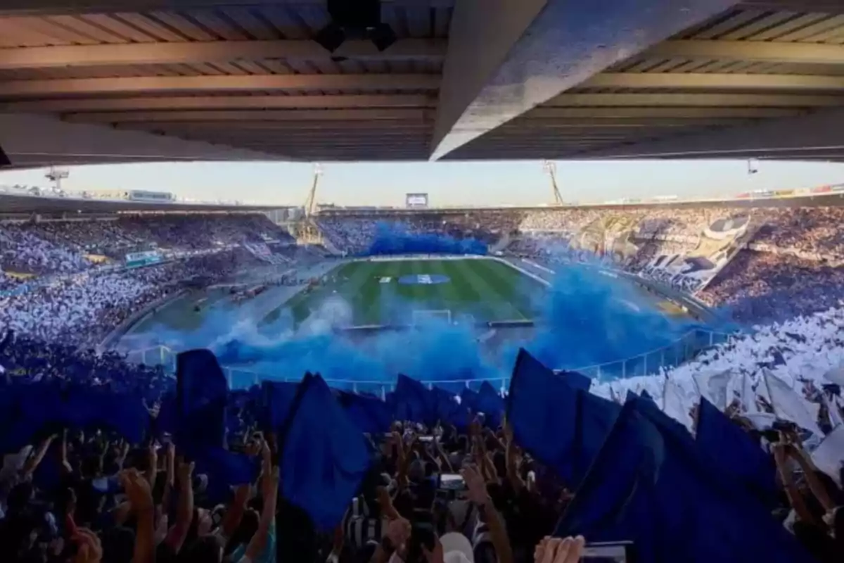 Aficionados en un estadio de fútbol ondeando banderas azules y blancas mientras se eleva humo azul sobre la cancha