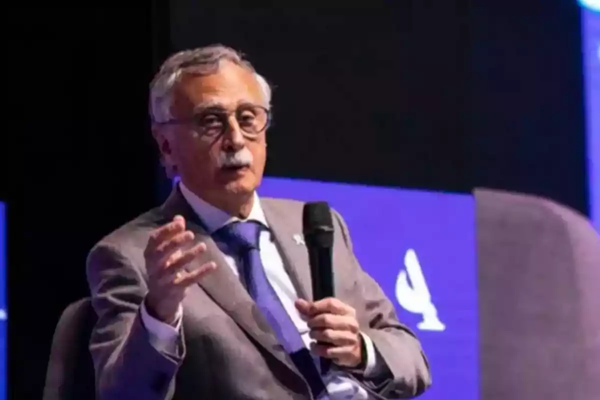 Gray-haired man with glasses speaking at an event while holding a microphone and wearing a gray suit with a blue tie Gray-haired man with glasses speaking at an event while holding a microphone and wearing a gray suit with a blue tie