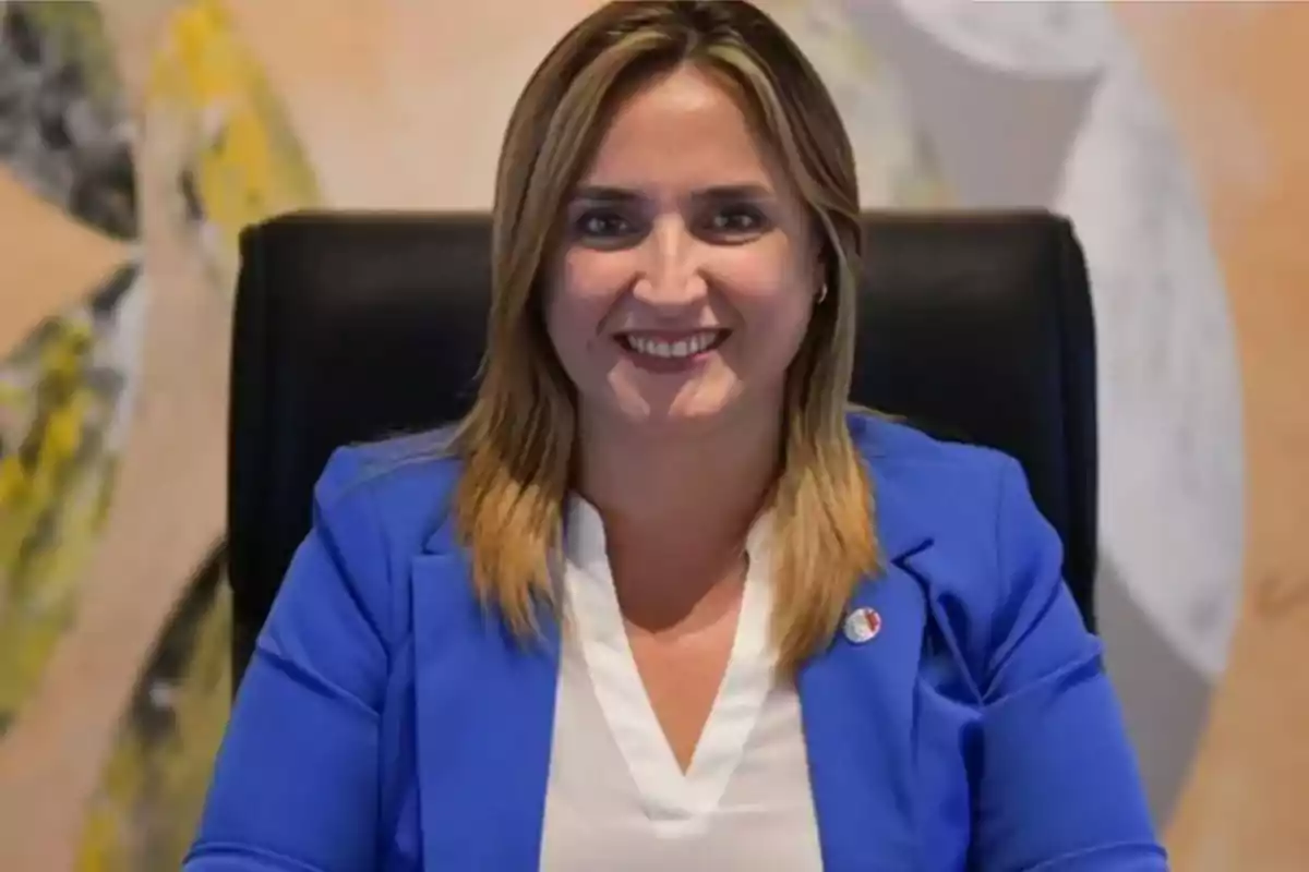 Mujer sentada en una silla negra vistiendo saco azul y blusa blanca sonriendo frente a un fondo abstracto. Mujer sentada en una silla negra vistiendo saco azul y blusa blanca sonriendo frente a un fondo abstracto.