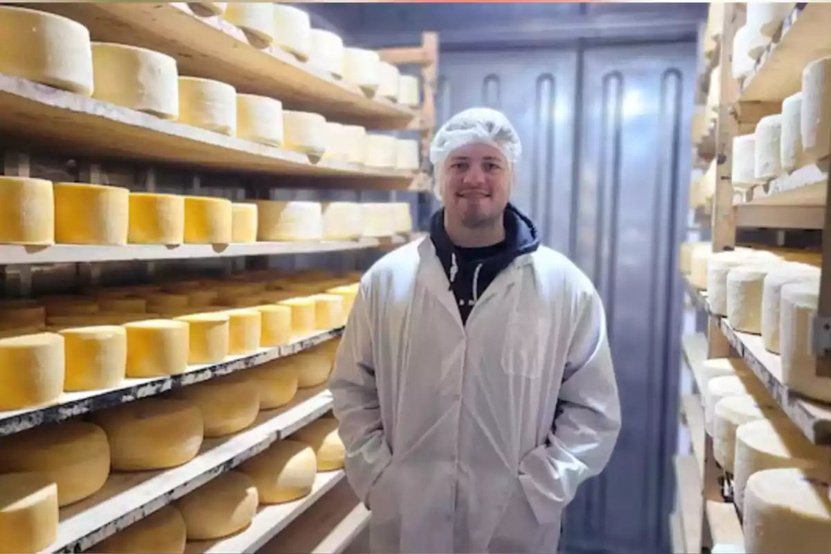 Hombre con bata y gorro de protección sonríe en una cámara de maduración rodeado de ruedas de queso en estantes de madera Hombre con bata y gorro de protección sonríe en una cámara de maduración rodeado de ruedas de queso en estantes de madera