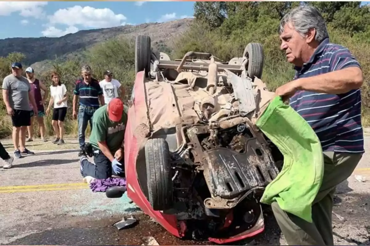 Un auto volcado sobre el asfalto mientras varias personas observan y un hombre sostiene una manta verde Un auto volcado sobre el asfalto mientras varias personas observan y un hombre sostiene una manta verde