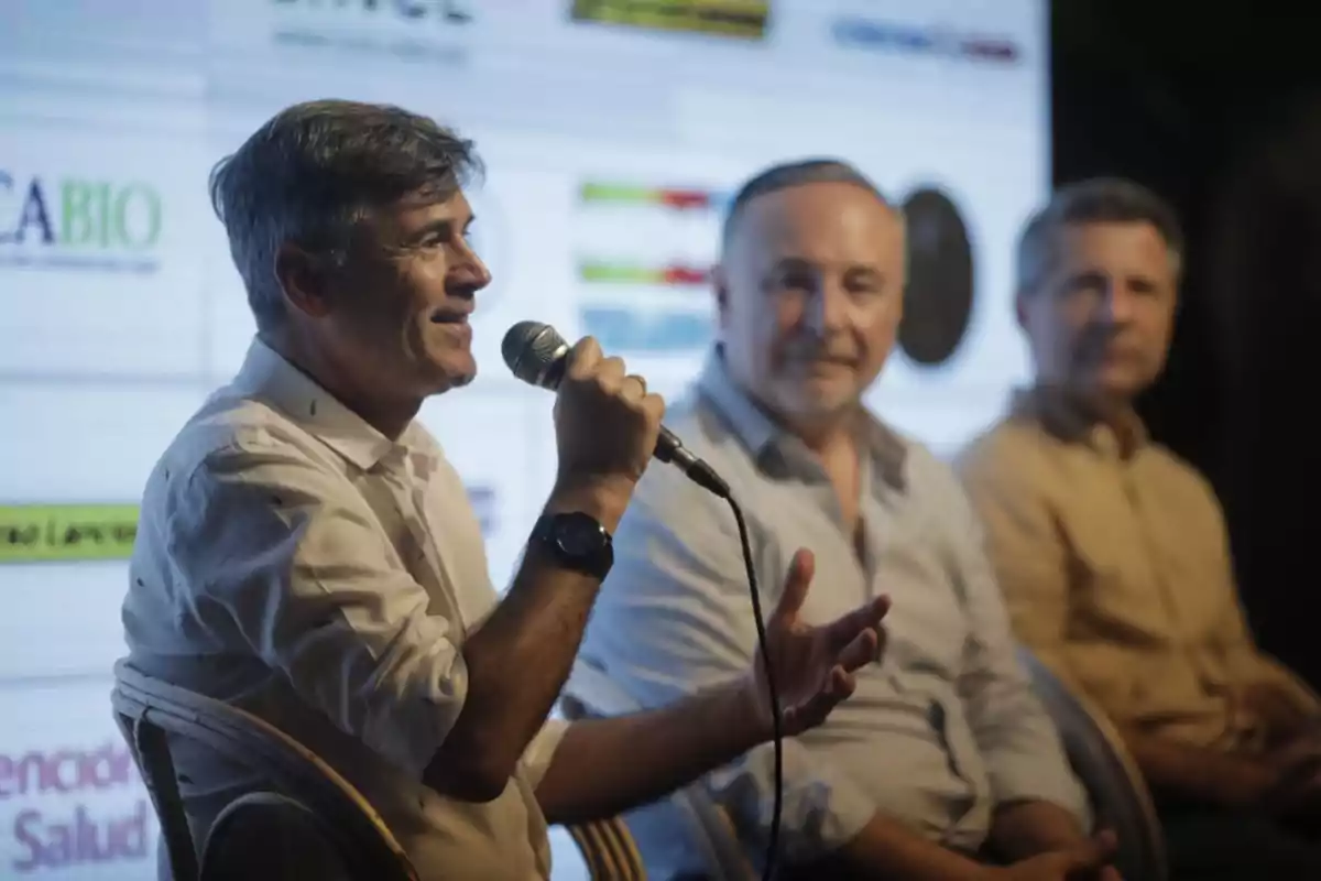 Man speaking into a microphone while two other men listen to him seated on a panel in front of a screen with logos. Man speaking into a microphone while two other men listen to him seated on a panel in front of a screen with logos.