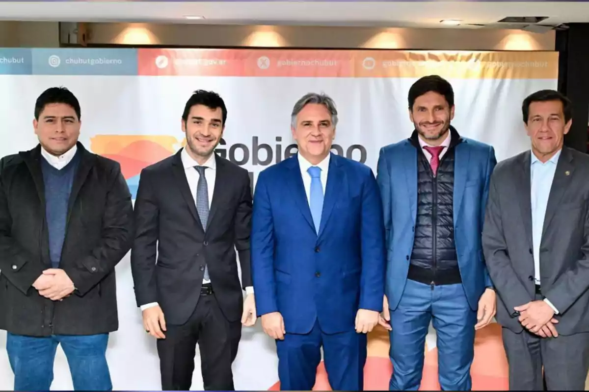 Five men pose together in front of an official government sign from Chubut, all dressed in formal attire and smiling at the camera. Five men pose together in front of an official government sign from Chubut, all dressed in formal attire and smiling at the camera.