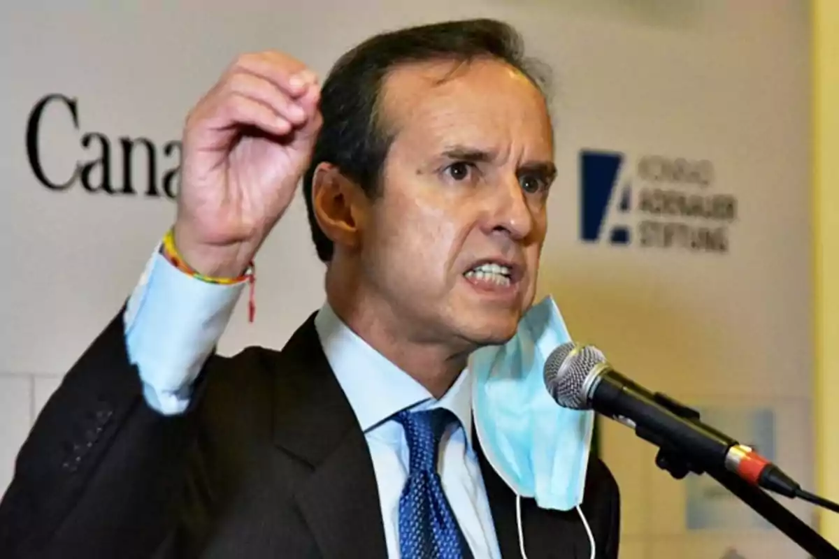 Man in a suit speaking into a microphone with a mask hanging from one ear and gesturing with his hand Man in a suit speaking into a microphone with a mask hanging from one ear and gesturing with his hand