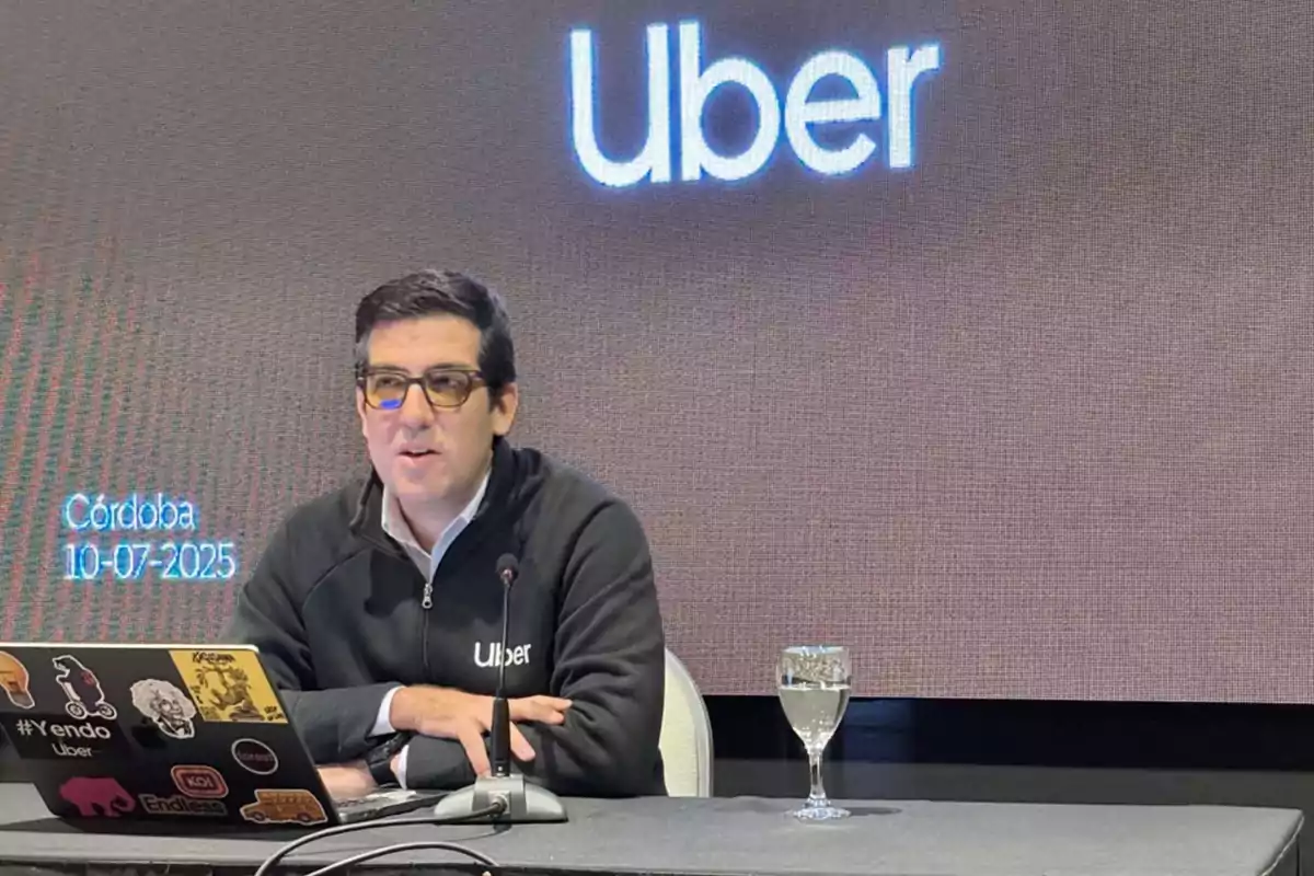 Hombre con gafas y chaqueta de Uber hablando en una conferencia con una laptop decorada con calcomanías y una copa de agua sobre la mesa, fondo con el logo de Uber y la palabra Córdoba junto a una fecha Hombre con gafas y chaqueta de Uber hablando en una conferencia con una laptop decorada con calcomanías y una copa de agua sobre la mesa, fondo con el logo de Uber y la palabra Córdoba junto a una fecha