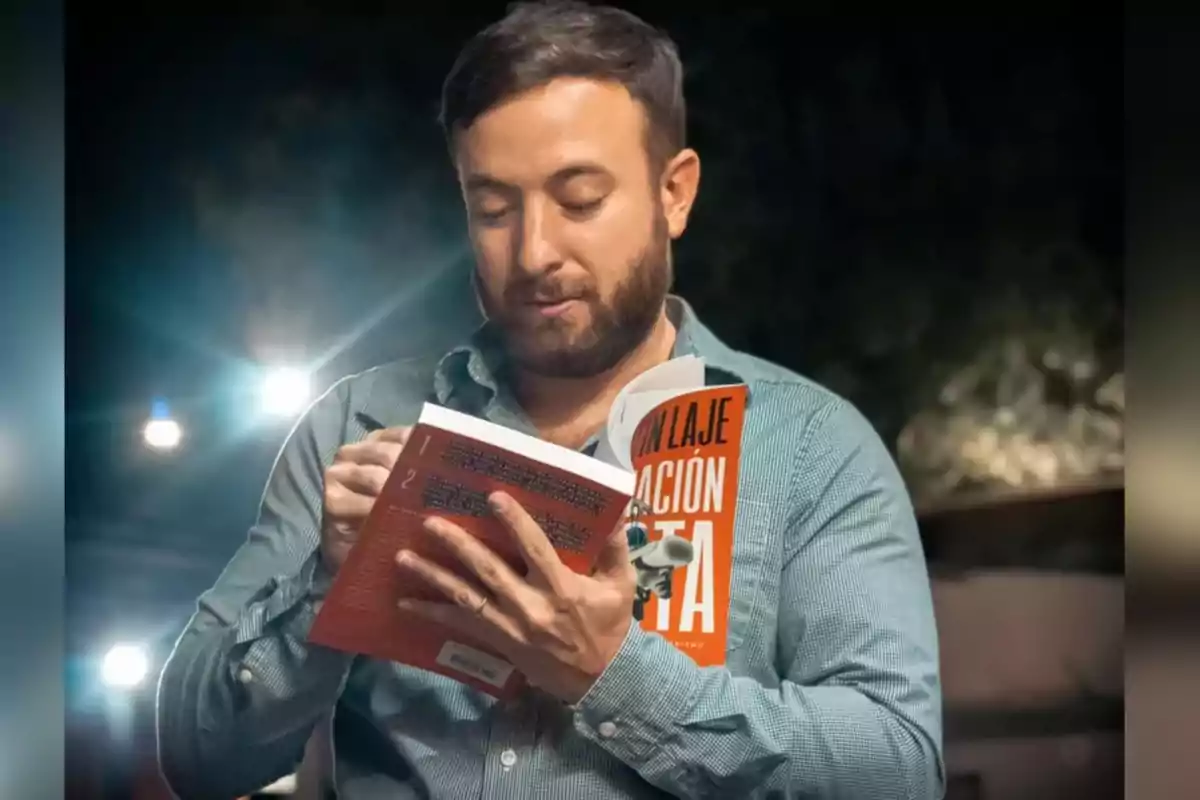 Bearded man reading a book with an orange cover while holding a cup in his other hand in a nighttime setting with lights in the background Bearded man reading a book with an orange cover while holding a cup in his other hand in a nighttime setting with lights in the background