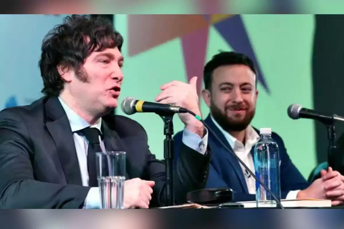 Two men in suits are sitting at a table during a conference; one of them is speaking into a microphone while the other smiles. Two men in suits are sitting at a table during a conference; one of them is speaking into a microphone while the other smiles.