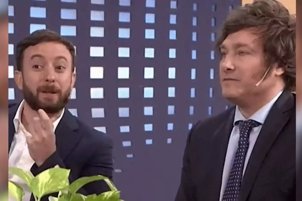Two men in suits are sitting on a television set; one of them is gesturing with his hand while the other keeps a serious expression. Two men in suits are sitting on a television set; one of them is gesturing with his hand while the other keeps a serious expression.
