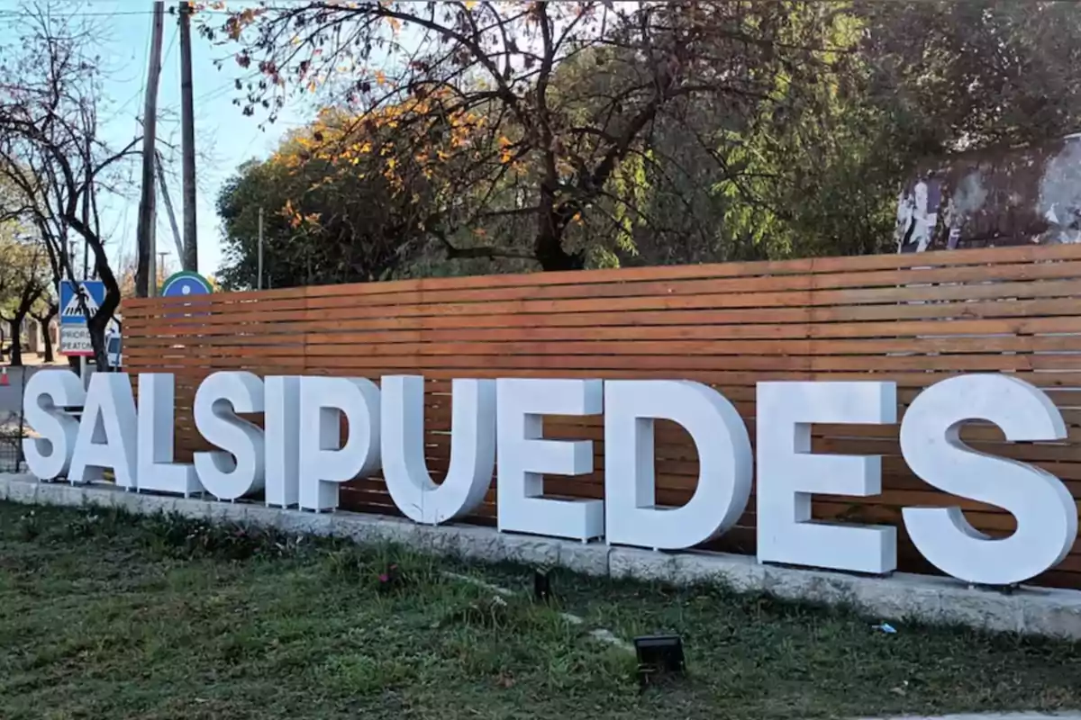 Letras blancas grandes que forman la palabra Salsipuedes frente a una cerca de madera y árboles al fondo Letras blancas grandes que forman la palabra Salsipuedes frente a una cerca de madera y árboles al fondo