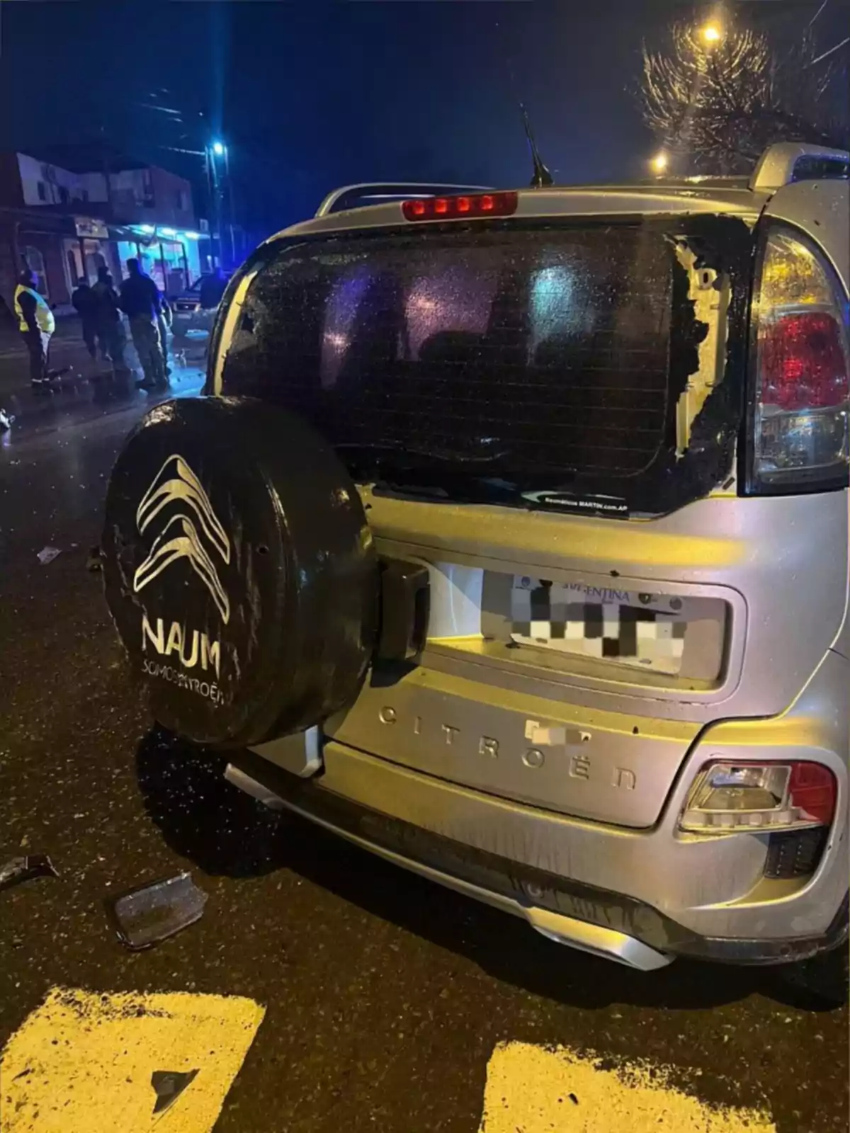 Auto Citroën plateado con la luneta trasera rota estacionado en una calle mojada de noche con personas y luces al fondo Auto Citroën plateado con la luneta trasera rota estacionado en una calle mojada de noche con personas y luces al fondo