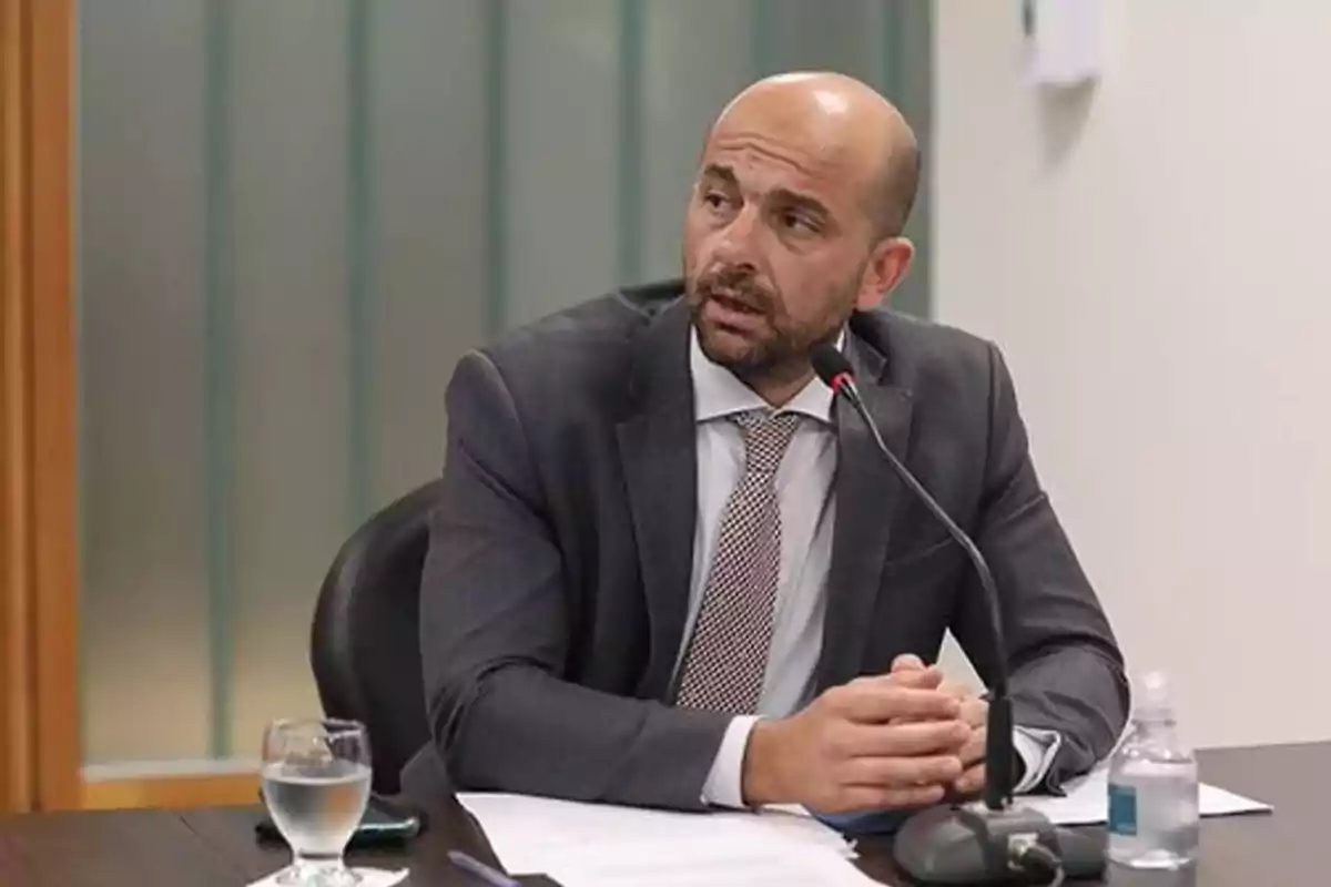 Hombre calvo con barba usando traje y corbata hablando en un micrófono durante una reunión en una oficina Hombre calvo con barba usando traje y corbata hablando en un micrófono durante una reunión en una oficina