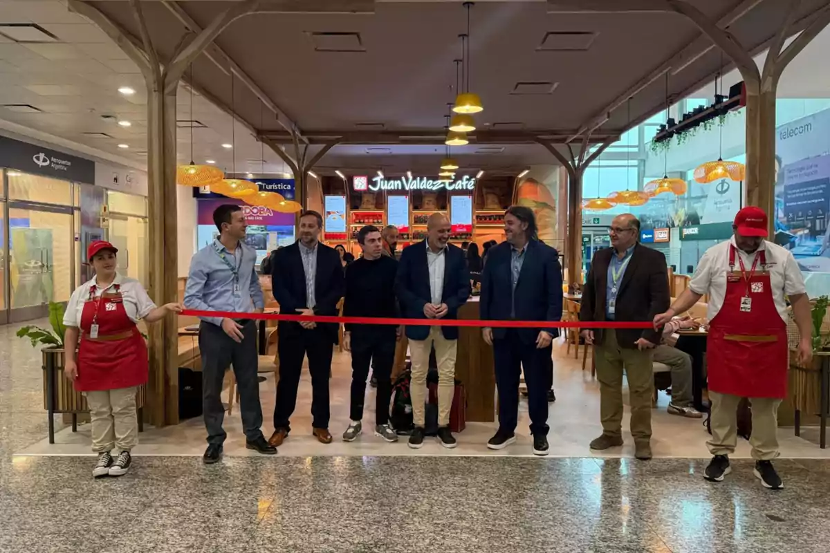 Varias personas participan en una ceremonia de inauguración frente a una cafetería Juan Valdez Café dentro de un centro comercial o aeropuerto, sosteniendo una cinta roja lista para ser cortada. Varias personas participan en una ceremonia de inauguración frente a una cafetería Juan Valdez Café dentro de un centro comercial o aeropuerto, sosteniendo una cinta roja lista para ser cortada.