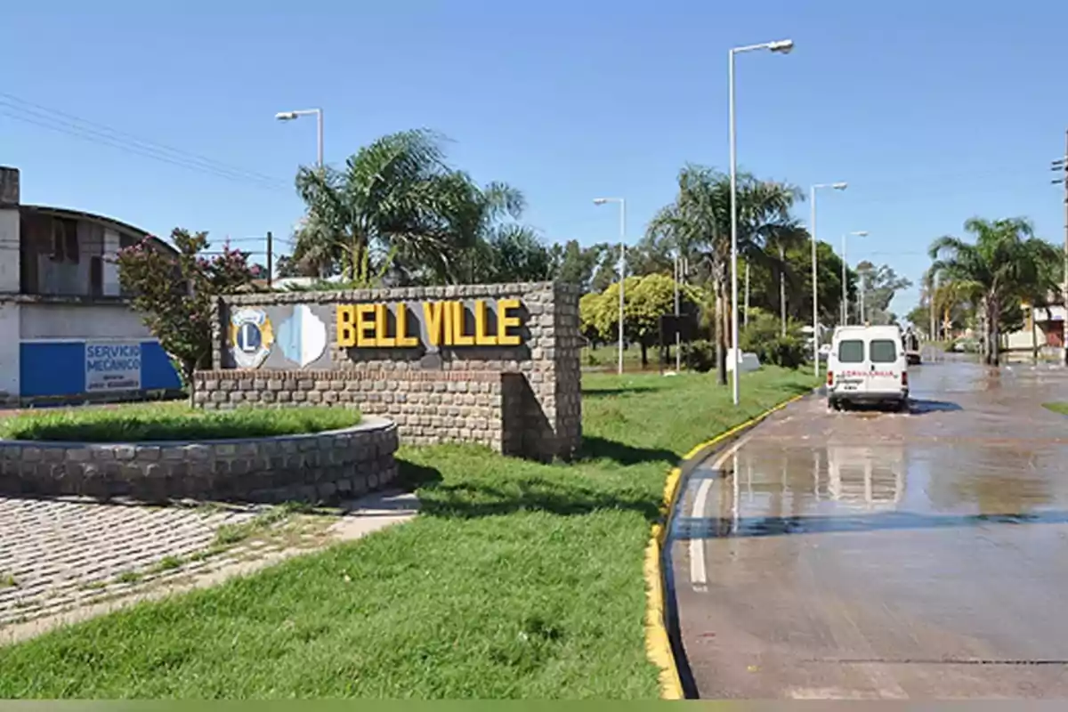 Cartel de bienvenida a Bell Ville junto a una calle con palmeras y una camioneta blanca en un día soleado