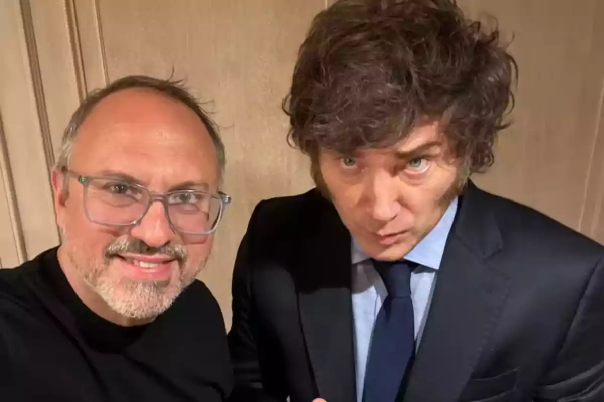 Dos hombres posan para una selfie, uno lleva gafas y camiseta negra, el otro viste traje oscuro y corbata azul Dos hombres posan para una selfie, uno lleva gafas y camiseta negra, el otro viste traje oscuro y corbata azul
