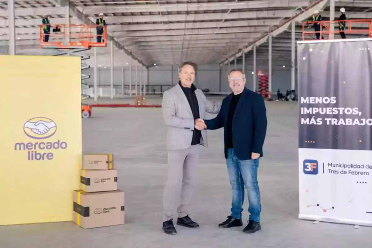 Dos hombres se dan la mano en el centro de un gran depósito en construcción, junto a cajas de Mercado Libre y un cartel que dice menos impuestos más trabajo de la Municipalidad de Tres de Febrero, mientras trabajadores con casco están en plataformas elevadoras al fondo. Dos hombres se dan la mano en el centro de un gran depósito en construcción, junto a cajas de Mercado Libre y un cartel que dice menos impuestos más trabajo de la Municipalidad de Tres de Febrero, mientras trabajadores con casco están en plataformas elevadoras al fondo.