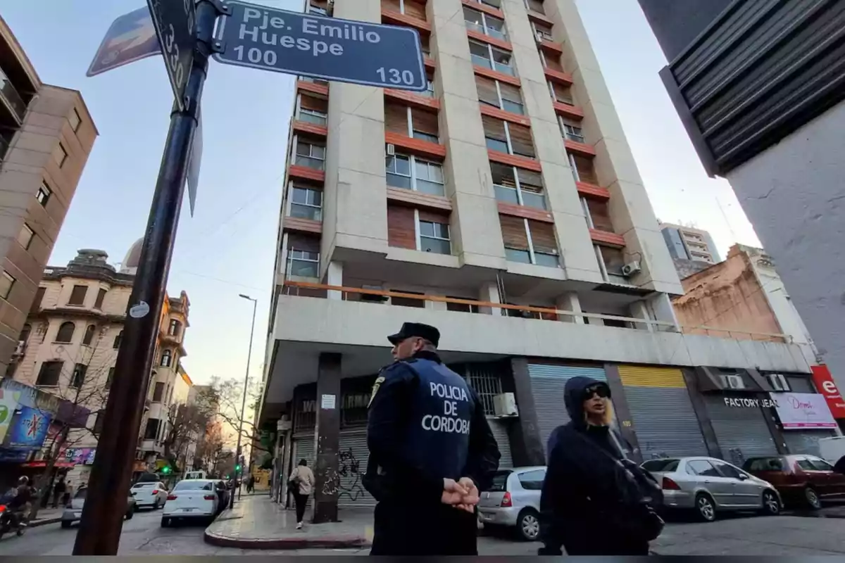 A police officer and a woman walk down an urban street next to a tall building and a sign that reads "Pje. Emilio Huespe."