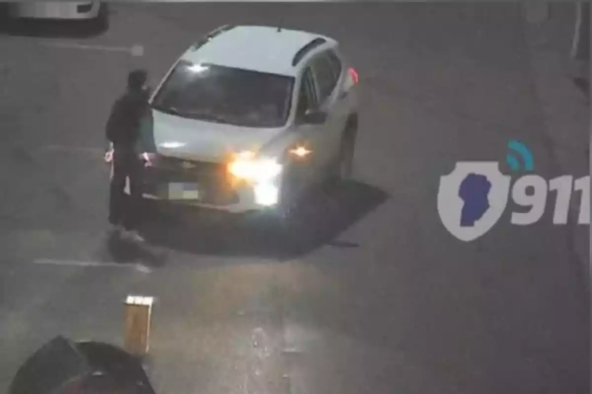 Person standing in front of a white car with its headlights on, on a street at night, and the 911 logo in the corner of the image Person standing in front of a white car with its headlights on, on a street at night, and the 911 logo in the corner of the image
