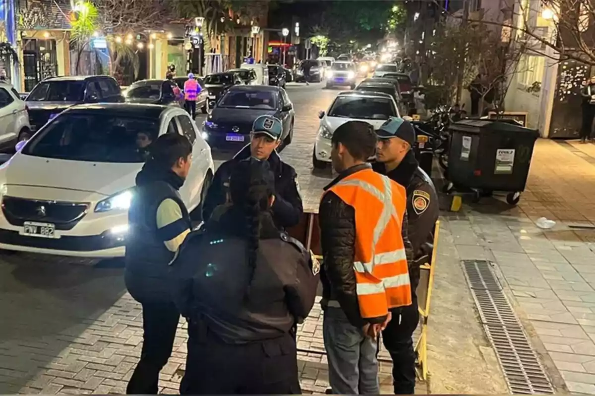 A group of police officers and a man wearing a reflective vest are talking on a busy street at night, with parked cars and lit-up storefronts in the background.