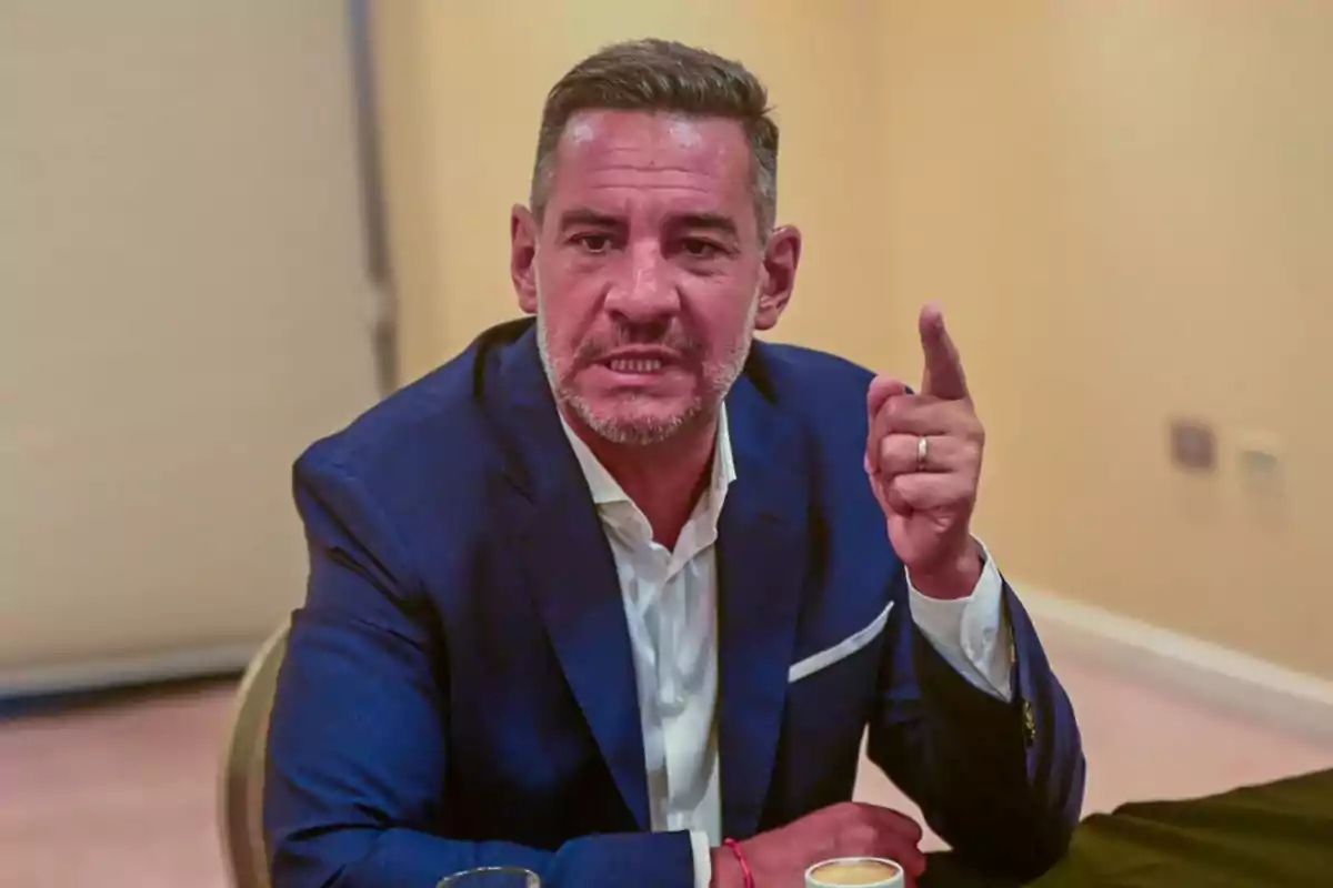 Short-haired, gray-bearded man wearing a blue jacket gesturing with his raised hand while seated at a table Short-haired, gray-bearded man wearing a blue jacket gesturing with his raised hand while seated at a table