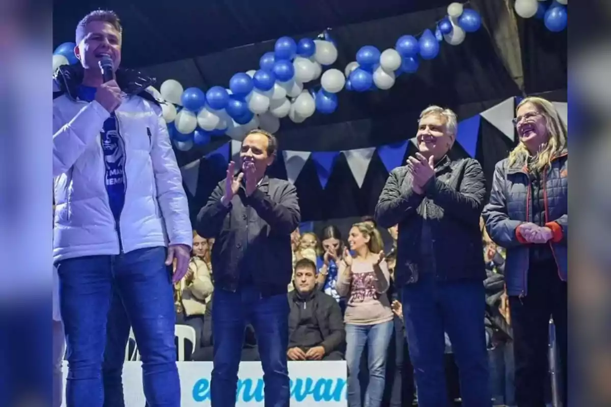 A man with a microphone speaks on a stage decorated with blue and white balloons and pennants while other people applaud and the audience watches in the background. A man with a microphone speaks on a stage decorated with blue and white balloons and pennants while other people applaud and the audience watches in the background.