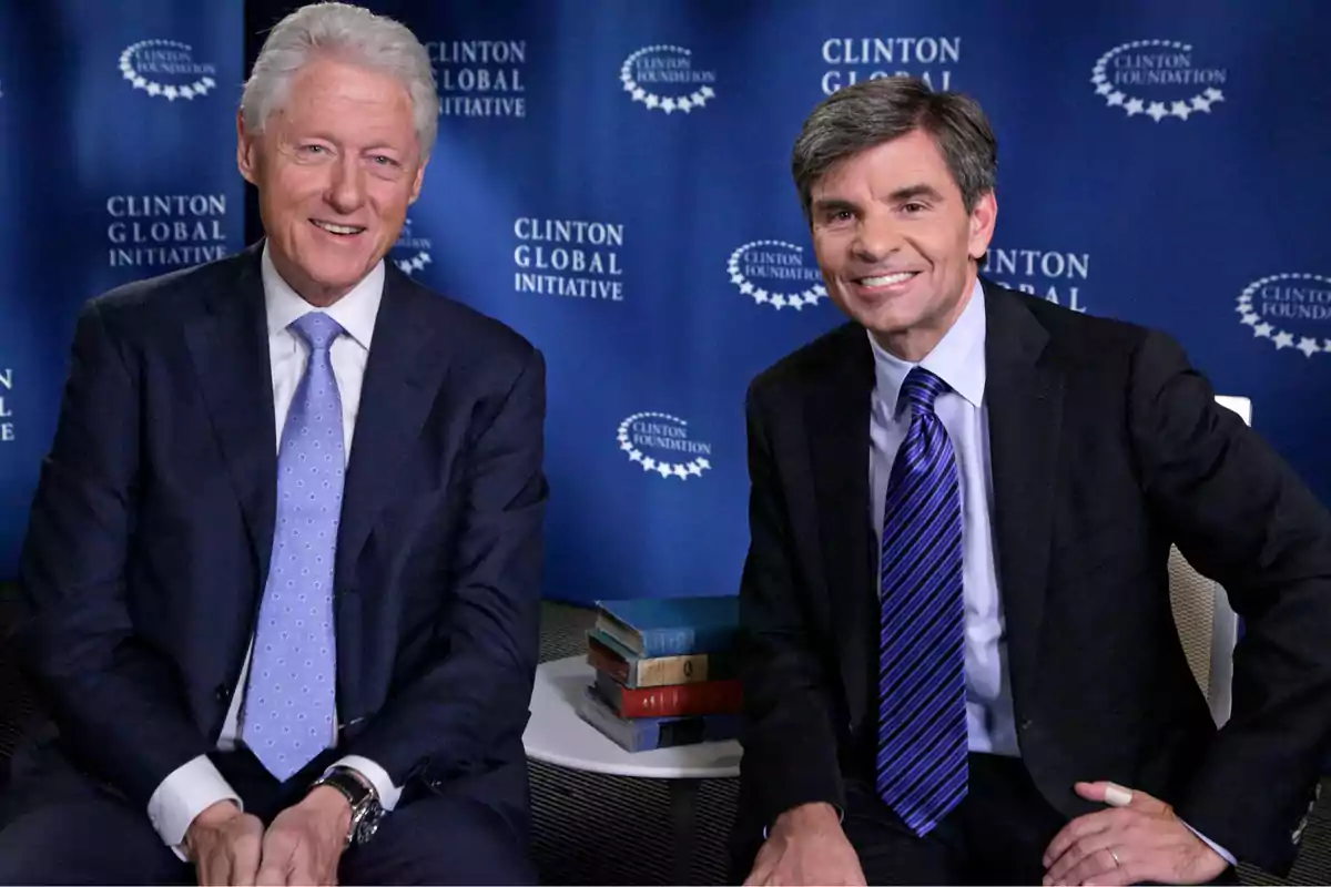 Dos hombres con traje y corbata sonríen sentados frente a un fondo azul con logotipos de la Clinton Foundation y Clinton Global Initiative, junto a una mesa con libros apilados. Dos hombres con traje y corbata sonríen sentados frente a un fondo azul con logotipos de la Clinton Foundation y Clinton Global Initiative, junto a una mesa con libros apilados.