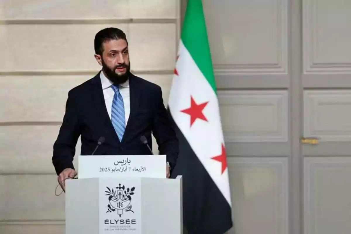 Un hombre con barba y traje oscuro habla en un podio con el logo del Palacio del Elíseo y una bandera con estrellas rojas y fondo verde, blanco y negro al fondo Un hombre con barba y traje oscuro habla en un podio con el logo del Palacio del Elíseo y una bandera con estrellas rojas y fondo verde, blanco y negro al fondo