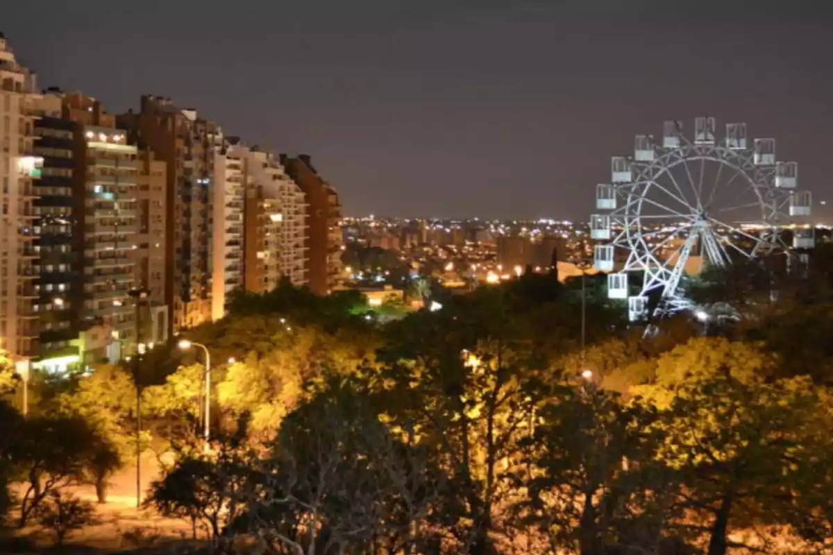 Vista nocturna de una ciudad con edificios altos a la izquierda, una rueda de la fortuna iluminada a la derecha y árboles en primer plano