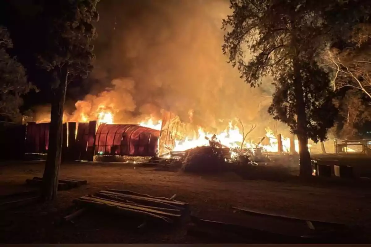Un edificio de madera arde intensamente en medio de la noche rodeado de árboles y humo denso Un edificio de madera arde intensamente en medio de la noche rodeado de árboles y humo denso