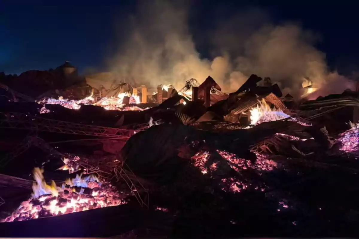 Restos de una estructura metálica y escombros en llamas con humo denso elevándose en la noche