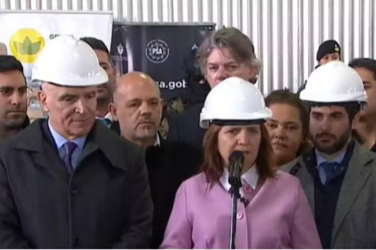 Varias personas con cascos de seguridad participan en un evento formal en un entorno industrial, una mujer habla frente a un micrófono mientras los demás observan. Varias personas con cascos de seguridad participan en un evento formal en un entorno industrial, una mujer habla frente a un micrófono mientras los demás observan.