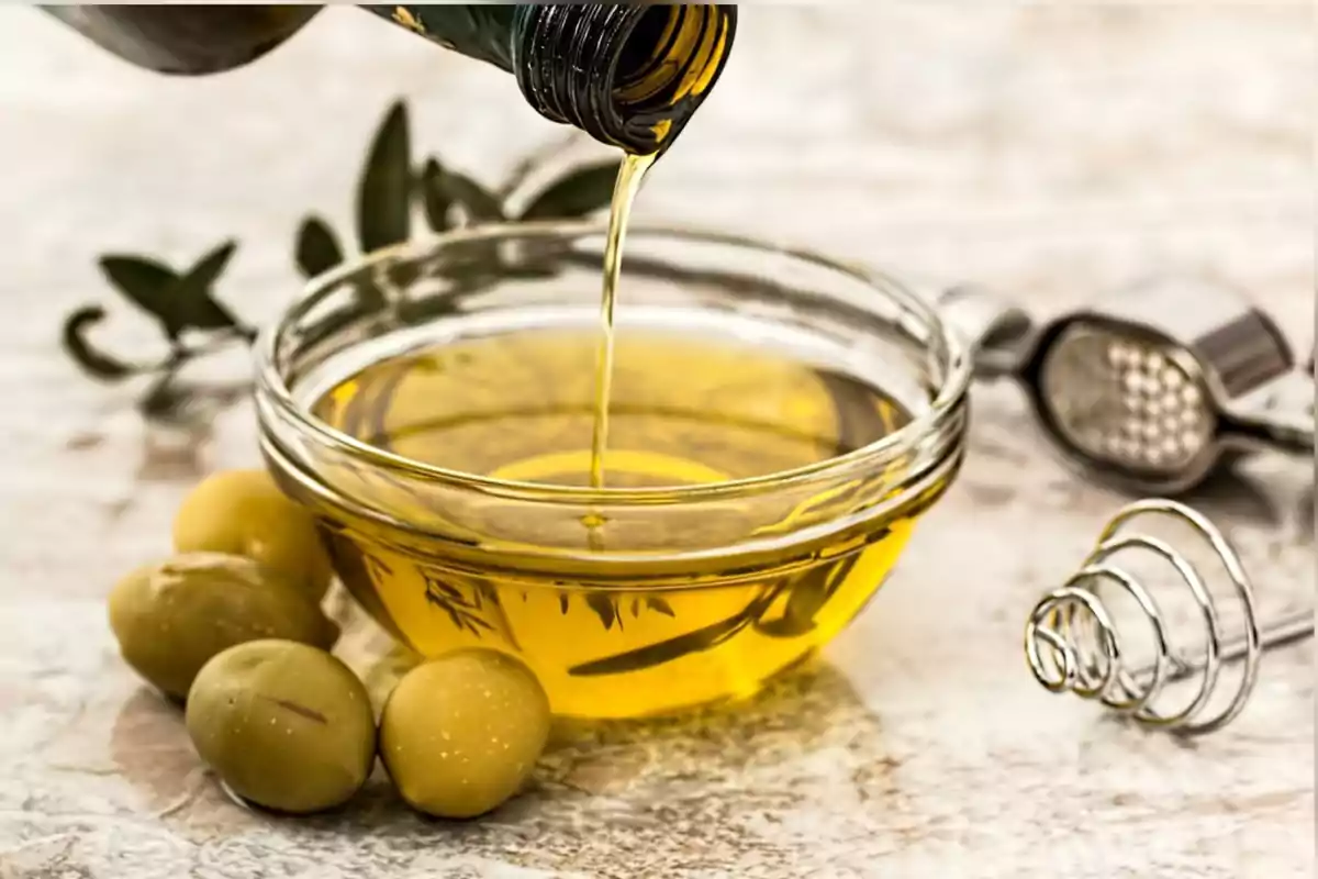 Olive oil being poured into a glass container next to green olives and kitchen utensils on a light-colored table