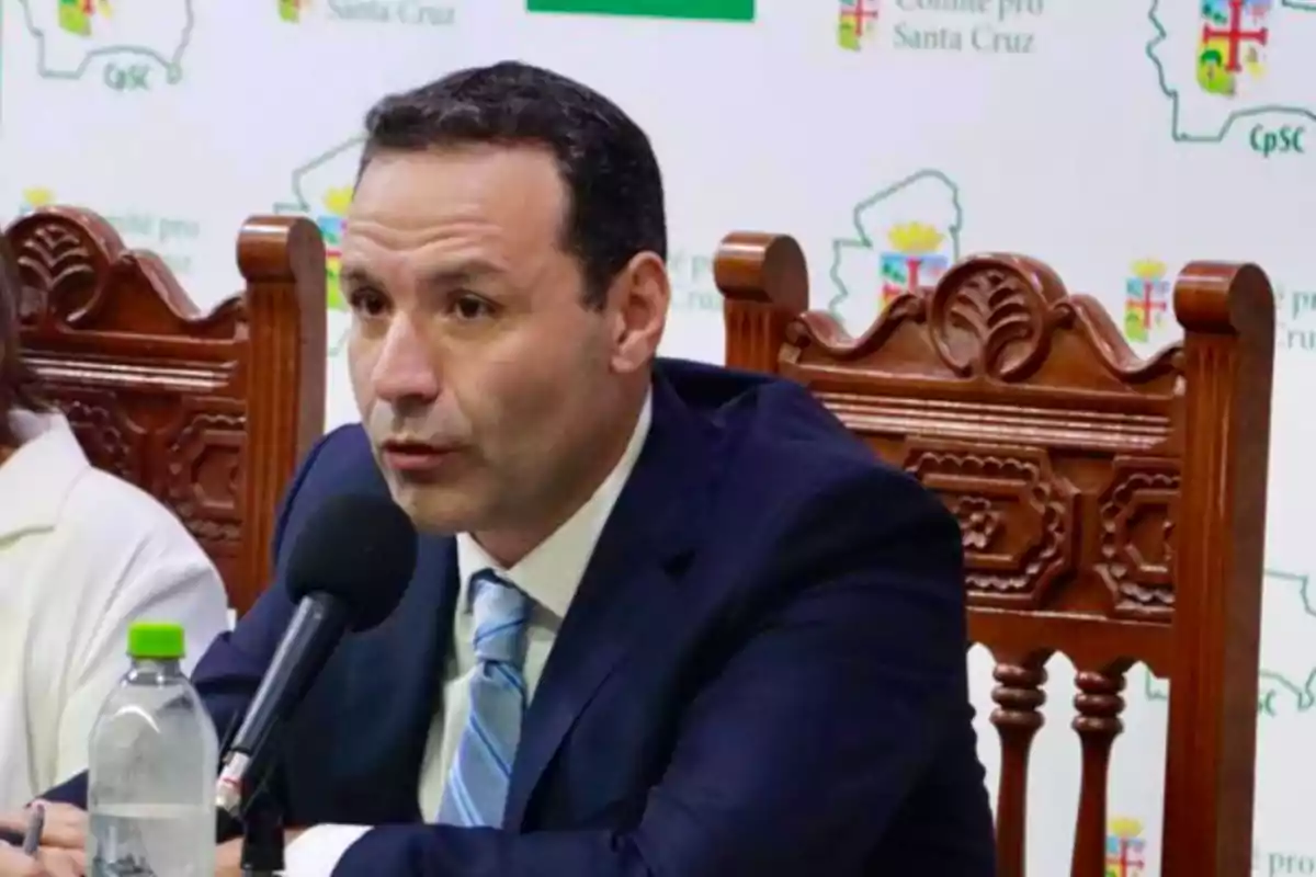 Hombre de traje azul hablando en una conferencia de prensa con un micrófono frente a él y una botella de agua sobre la mesa Hombre de traje azul hablando en una conferencia de prensa con un micrófono frente a él y una botella de agua sobre la mesa