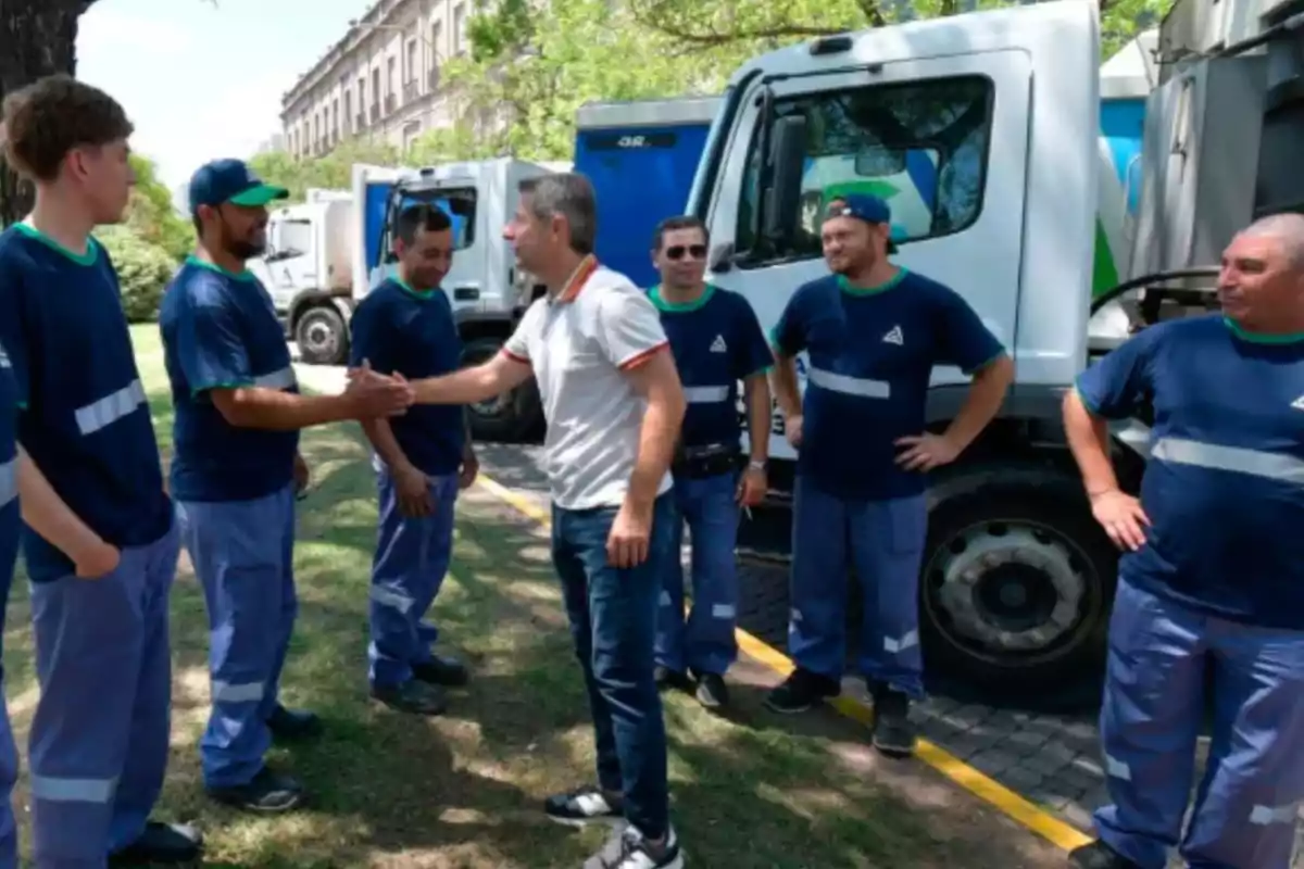 Un grupo de trabajadores con uniforme azul está de pie junto a camiones de basura mientras uno de ellos saluda de mano a un hombre vestido de civil en un entorno al aire libre
