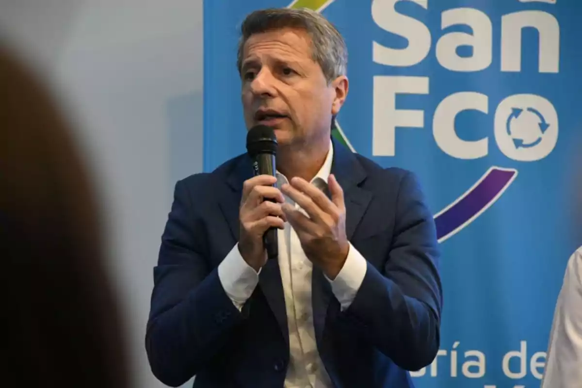 Hombre de cabello canoso con saco azul y camisa blanca hablando por micrófono frente a un cartel azul que dice San Fco Hombre de cabello canoso con saco azul y camisa blanca hablando por micrófono frente a un cartel azul que dice San Fco