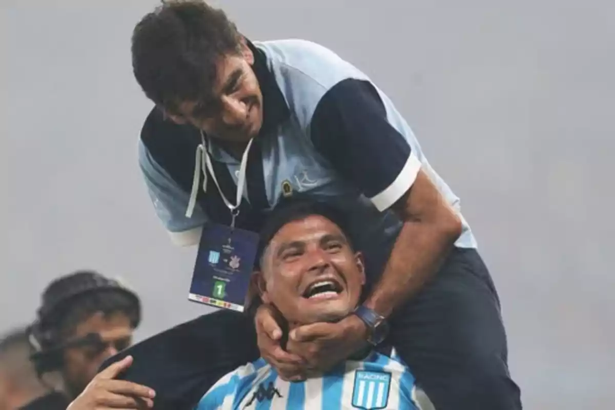 Two men celebrate enthusiastically in a stadium; one of them, wearing a Racing Club uniform, smiles while the other hugs him from behind around the neck. Two men celebrate enthusiastically in a stadium; one of them, wearing a Racing Club uniform, smiles while the other hugs him from behind around the neck.