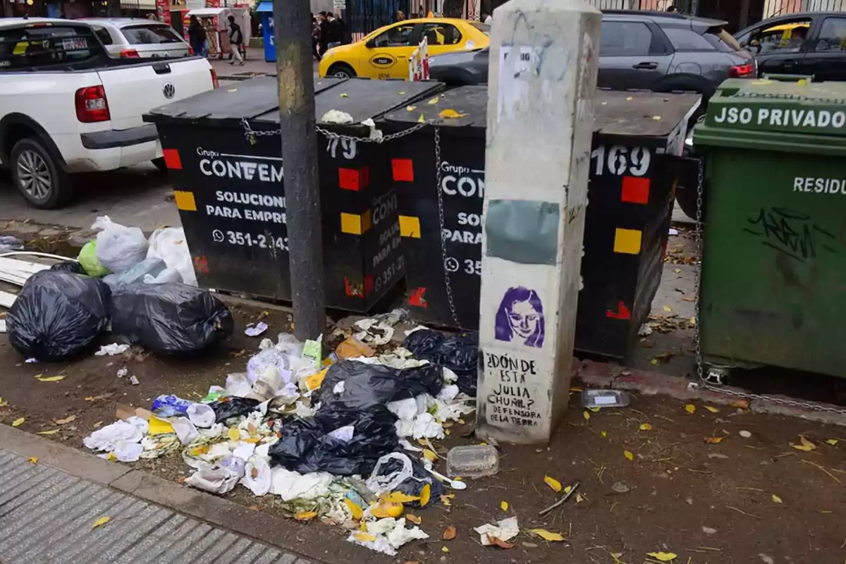 Basura y bolsas de residuos acumuladas en la vereda junto a contenedores de basura y un poste con grafiti en una calle urbana Basura y bolsas de residuos acumuladas en la vereda junto a contenedores de basura y un poste con grafiti en una calle urbana